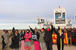 An image taken of protestors at Standing Rock. Several people hold signs, two of which say "water is life" and "defend the sacred."