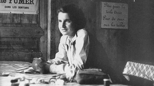 Black and white photograph of Rosalind Franklin sitting at a table, looking to the side with a thoughtful expression.