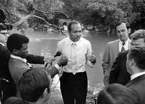Carl Stokes standing in front of the Cuyahoga river talking to a group of reporters